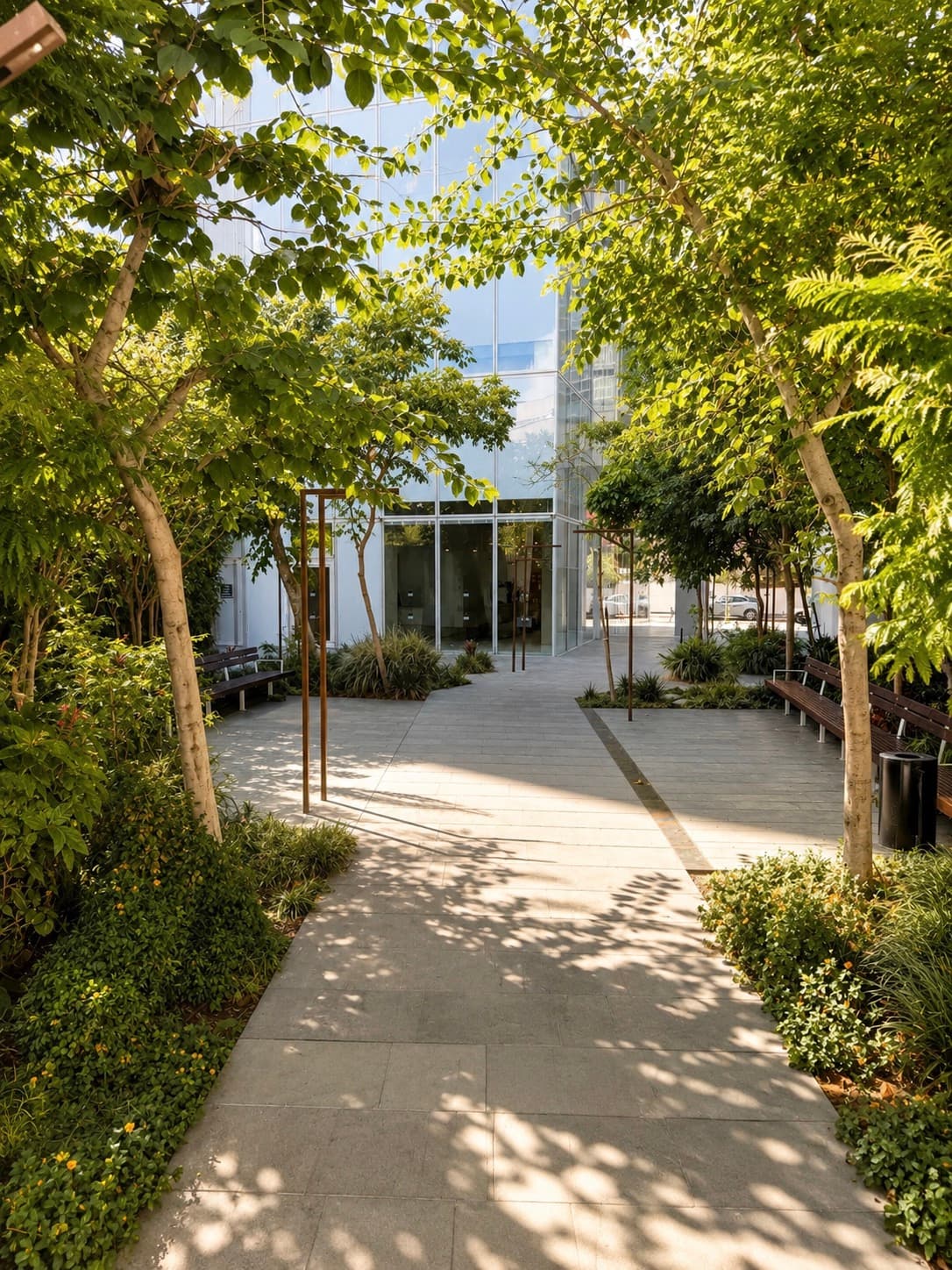 Allée dallée bordée d'arbres et de bancs, menant à l'entrée vitrée de l'immeuble.