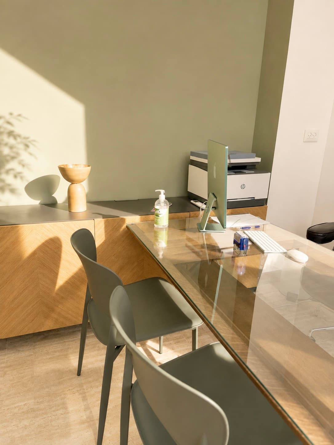 Plan de bureau en verre, deux chaises, écran d'ordinateur et imprimante sur un meuble bas, mur vert olive.