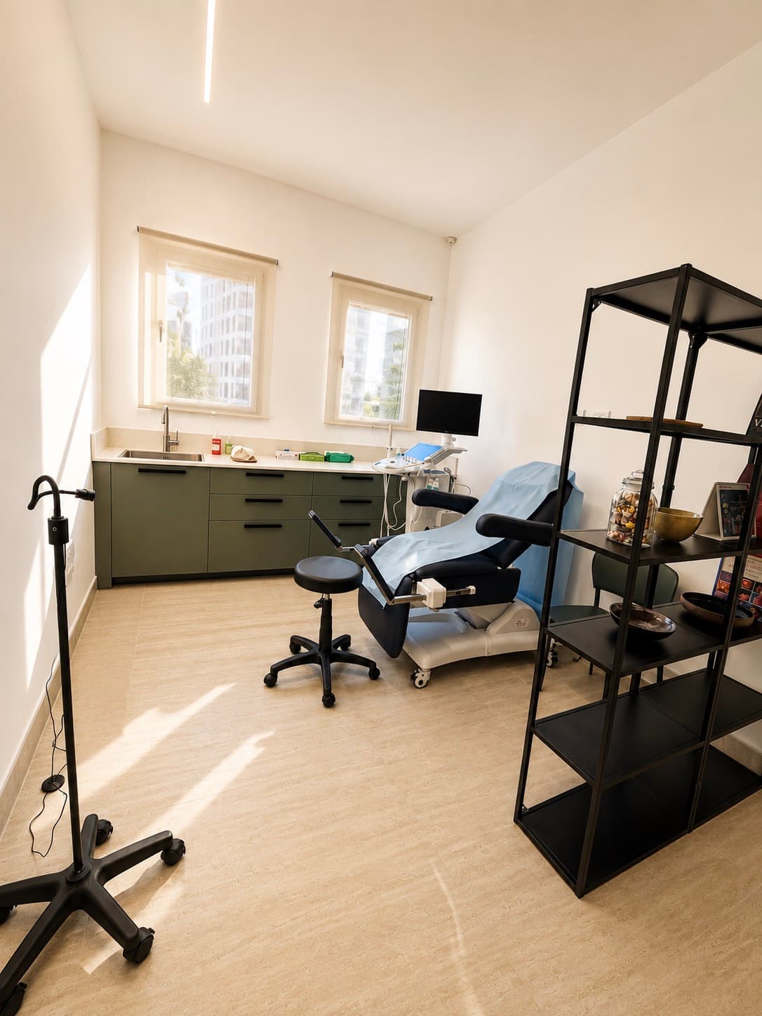 Salle d'examen : fauteuil gynécologique recouvert d'un drap bleu, échographe, meuble vert olive et deux fenêtres.